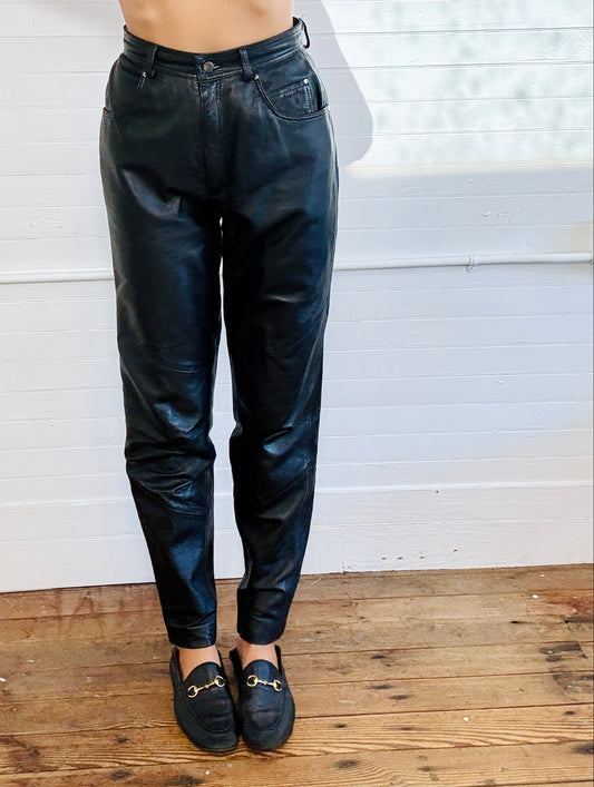 A person wearing shiny black tapered pants and black loafers, standing on a wooden floor against a white paneled wall.
